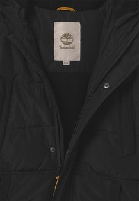 Giacca nera imbottita con zip gialla, dotata di foderatura in pile e di un'etichetta con il logo Timberland. Visibili le chiusure a zip e a scatto.