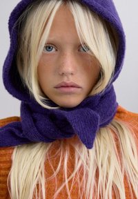 Capucha y bufanda de punto morado, textura suave. Cabello rubio claro con una ligera onda, enmarcado por la bufanda. Piel pálida con pecas.