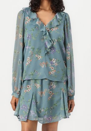 Kvinde iført en blå blomsterlangærmet bluse med flæset halsudskæring og matchende plisseret nederdel mod en ensfarvet hvid baggrund.