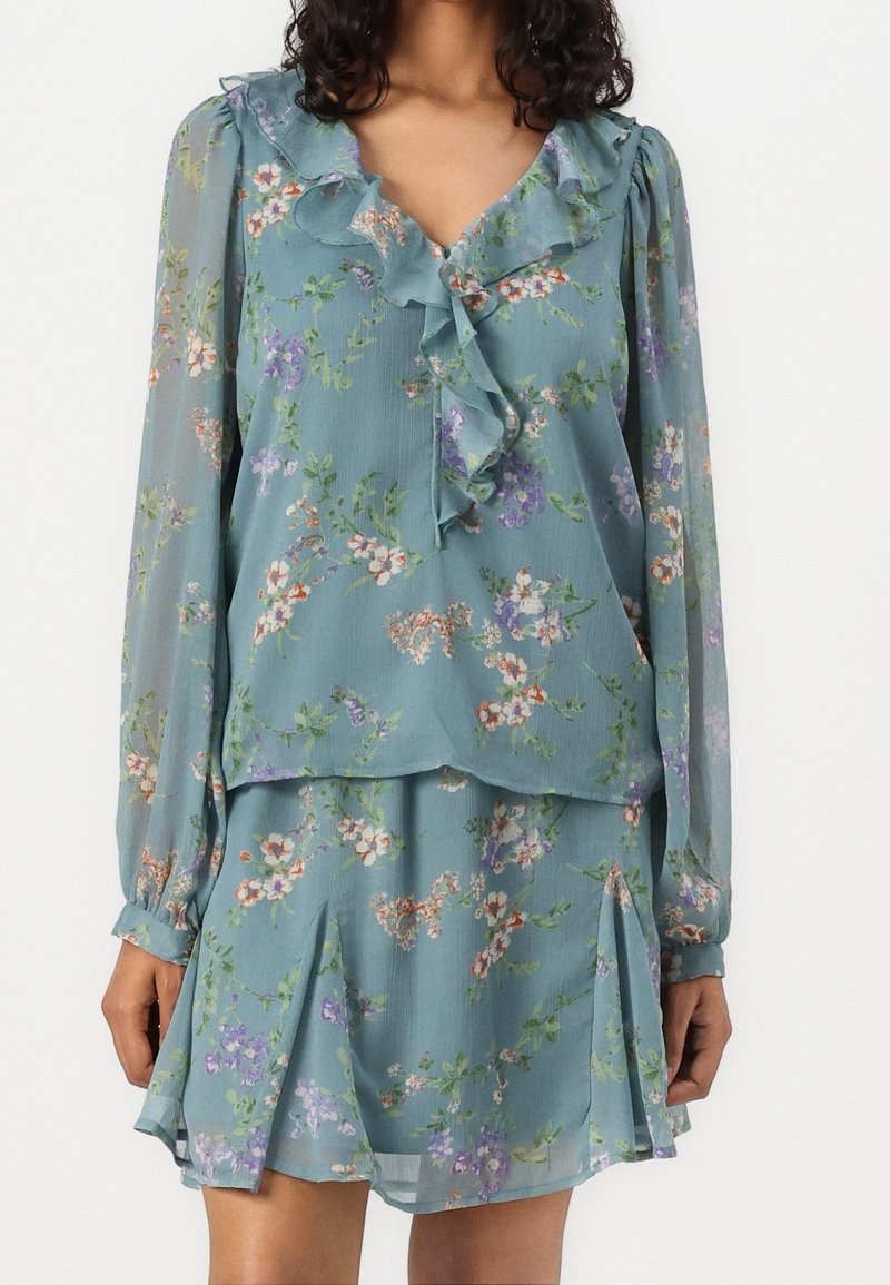 Femme portant un chemisier bleu à manches longues à motif floral avec un col volanté et une jupe plissée assortie sur un fond blanc uni.