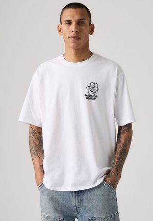 Junger Mann mit kurzen Haaren und Tattoos, der ein weißes T-Shirt mit einer kleinen Grafik und dem Text "Made for Workin'" auf der Brusttasche trägt.