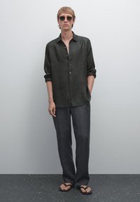 Donkergroen linnen shirt met knopen, gecombineerd met donkere gestreepte broek en zwarte slippers; model draagt ronde zonnebril.