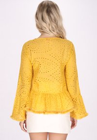 Gelbes Peplum-Oberteil aus strukturiertem Stoff, mit Ausschnittmustern, langen Glockenärmeln und Fransenakzenten entlang dem Saum und den Ärmeln.