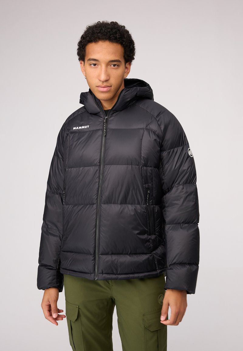 La veste noire rembourrée présente un avant zippé, une capuche et des poches latérales. Le matériau est lisse et brillant avec un motif cousu. Logo sur la poitrine.