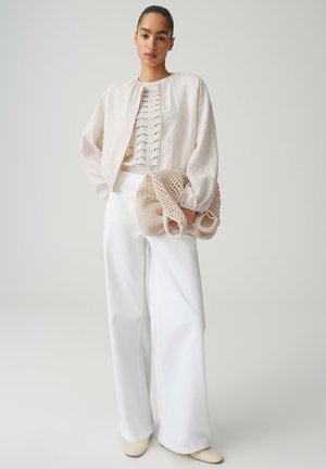 Frau trägt einen cremefarbenen Strickpullover, eine hellbeige Jacke, weiße Hosen mit weitem Bein und hält eine beigetone Häkel-Tasche vor hellem einfarbigem Hintergrund.