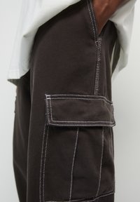 Mörkbruna cargobyxor med vit sömnad, sidoflappficka och texturerad tyg, kombinerat med en ljusfärgad t-shirt.
