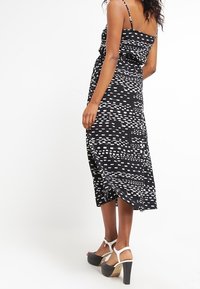 Woman wearing a black midi dress with white abstract patterns and white high-heeled sandals, facing away against a white background.