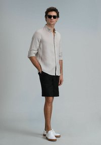 Lichtgekleurde stof shirt met opgerolde mouwen, zwarte shorts en witte sneakers met bruine zolen. Model met donkere zonnebril, rechtopstaand.