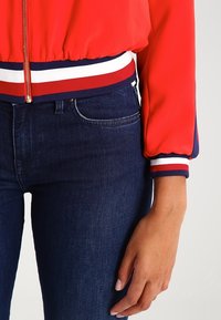Röd bomberjacka med elastiska muddar med blå och vita ränder. Kombinerad med mörkblå jeans. Slätt tyg och figurnära design.