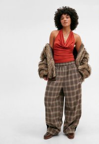 Mulher com cabelo encaracolado vestindo uma blusa halter vermelha, calças oversized de xadrez, sapatos castanhos e uma jaqueta de pele drapeada sobre os braços.