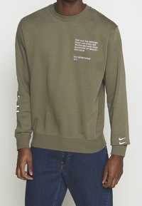 Olivově zelená mikina s bílým textem na přední straně a rukávu. Měkká látka, kulatý výstřih, žebrované manžety a vyšívané logo Nike na rukávu.
