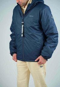 Veste isolante bleu marine avec capuche, dotée de poignets élastiques, d'une fermeture éclair à l'avant et d'une étiquette de logo. Associée à un pantalon de survêtement couleur crème.