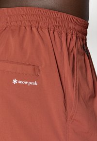 Rot-braune Shorts mit elastischem Bund, ausgestattet mit einer Gesäßtasche und einem weißen "Snow Peak"-Logo an der Seite. Glatte Stoffoberfläche.