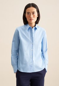 Camicia azzurro chiaro con colletto a punta, maniche lunghe e bottoni bianchi. Indossata con pantaloni blu navy scuri, caratterizzati da una texture liscia.