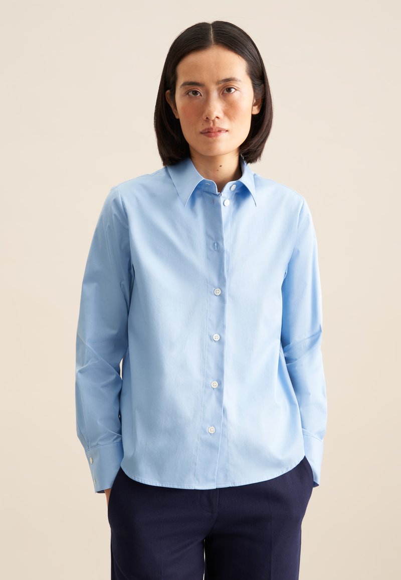 Camicia azzurro chiaro con colletto a punta, maniche lunghe e bottoni bianchi. Indossata con pantaloni blu navy scuri, caratterizzati da una texture liscia.