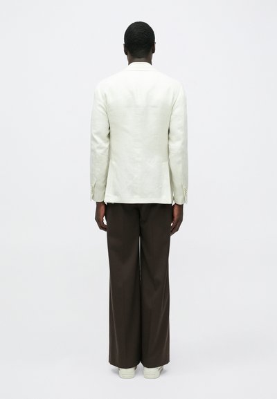 Homme debout, de dos, portant un blazer blanc, un pantalon large marron foncé et des baskets blanches, sur un fond clair uni.