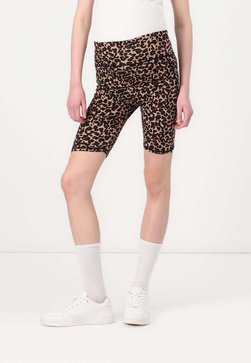 Personne portant un short de cycliste taille haute à imprimé léopard, des chaussettes blanches montantes et des baskets blanches, debout devant un fond clair uni.
