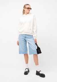 Felpa bianca con cappuccio, pantaloni corti in denim azzurro chiaro, scarpe platform nere e una piccola borsa nera. Gli accessori includono occhiali da sole in acetato tartarugato.