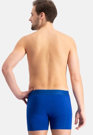 Mann mit dem Rücken zur Kamera, trägt blaue Bamboo Basics Boxershorts, rechte Hand in die Hüfte gestützt, vor schlichtem hellem Hintergrund.