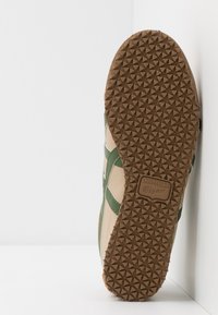 Baskets Onitsuka Tiger avec une semelle en caoutchouc marron texturé, un dessus crème et des accents verts. Présente un logo distinctif sur la semelle.