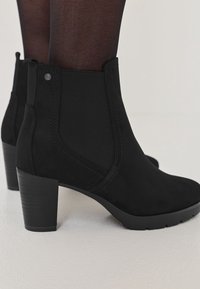 Femme portant des bottines en daim noir à talons carrés et des collants noirs transparents, debout sur une surface de couleur claire.