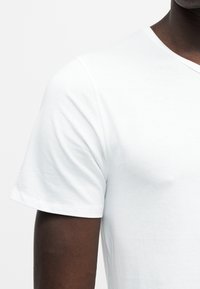 Nahaufnahme einer Person, die ein schlichtes weißes T-Shirt mit kurzen Ärmeln und Rundhalsausschnitt trägt und den Oberarm sowie die Schulter zeigt.