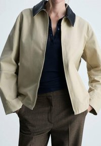 Veste beige avec un col noir, dotée d'une fermeture éclair à l'avant et de manches longues. Portée par-dessus un polo bleu marine et un pantalon marron, avec une texture lisse.
