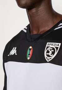 Maillot de sport noir et blanc avec un design en V, arborant le logo Kappa, le badge du club de Spezia et un emblème de drapeau italien sur la poitrine.