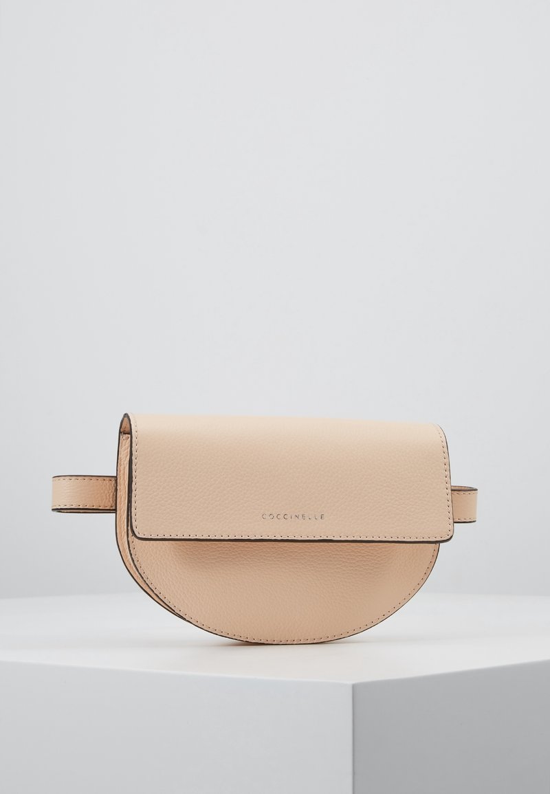 Sac banane en cuir beige en forme de demi-lune avec fermeture à rabat et coutures discrètes, posé sur un piédestal blanc sur fond uni.