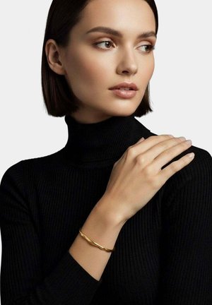 Donna con capelli corti e scuri, indossa un dolcevita nero, poggia la mano sulla spalla e porta un braccialetto d'oro intrecciato.