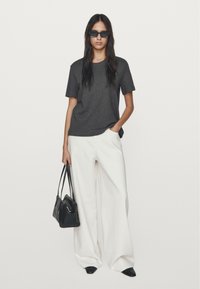 T-shirt grigio a maniche corte abbinato a pantaloni bianchi a gamba larga. Il modello tiene in mano una piccola borsa nera e indossa occhiali da sole neri.