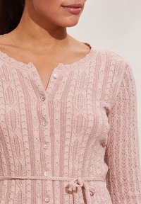 Jasnoróżowy sweter na drutach z fakturowanym wzorem, okrągłym, falowanym dekoltem oraz zapięciami na guziki z przodu, z paskiem do wiązania.