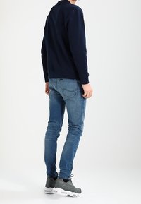 Navy sweatshirt, slim-fit blue jeans, and gray sneakers with white soles. Casual outfit displayed from a back view, featuring smooth textures.