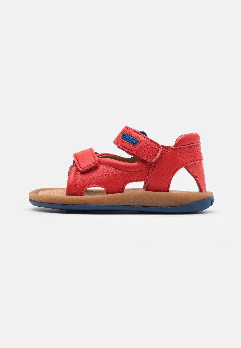 Sandale pour tout-petit en cuir rouge avec deux brides ajustables, semelle intérieure rembourrée couleur tan, et semelle extérieure souple bleu foncé sur fond blanc.