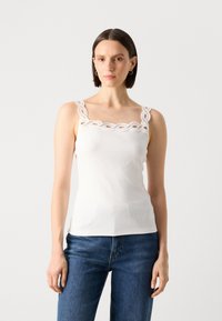 White ribbed tank top with decorative lace trim along the square neckline and adjustable straps, paired with blue denim jeans.