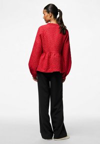 Sarkans teksturēts blūze ar pūkainām piedurknēm un sašaurinātu jostasvietu, apvienots ar melniem platām kājām biksēm un ikdienas apaviem, skatīts no aizmugures.