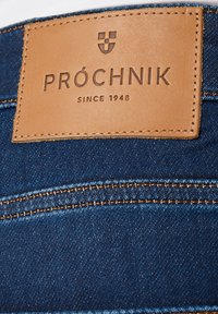 Brązowa skórzana metka na niebieskich dżinsach z wypisaną frazą "PROCHNIK SINCE 1948" oraz małym logo tarczy nad tekstem.