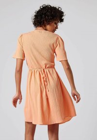 Perzikkleurige jurk met korte mouwen, van een textuurstof, met een getailleerde taille en een trekkoord aan de achterkant. De uitlopende rok valt boven de knie.