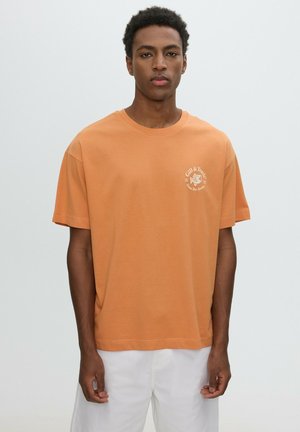 Joven con una camiseta naranja de manga corta con un pequeño logo blanco y pantalones blancos, de pie frente a un fondo claro y sencillo.