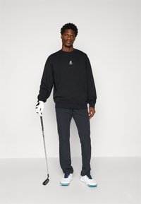 Melna džemperis ar baltu logo, tumši pelēkas bikses, baltas kedas ar zilo akcentu. Tur rokā golfa nūju, valkājot baltu cimdu.