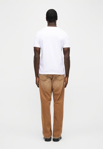 Άνδρας με την πλάτη γυρισμένη, φοράει ένα απλό λευκό μπλουζάκι, καφέ ξεθωριασμένα τζιν με ίσια ποδόγυρα και μαύρα παπούτσια, στέκεται μπροστά σε ένα απλό λευκό φόντο.