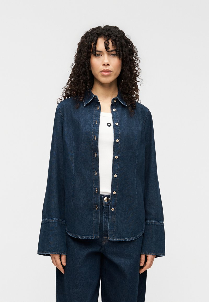 Πουκάμισο τζιν σε σκούρο μπλε, με κουμπιά μπροστά, μυτερό γιακά και φαρδιά μανίκια. Συνδυασμένο με λευκό τοπ και σκούρο τζιν.