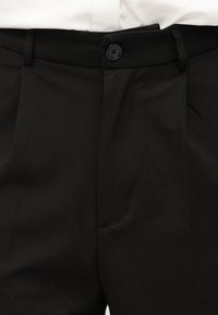 Κοντινό πλάνο μαύρου παντελονιού με κουμπωτή μέση και ορατές θηλιές ζώνης πάνω από ένα λευκό πουκάμισο.