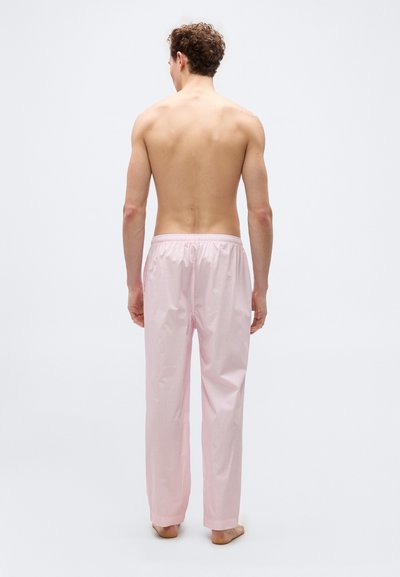 Άνδρας που στέκεται ξυπόλυτος, από πίσω, φορώντας φαρδιά πυτζάμα με ριγέ ανοιχτό ροζ παντελόνια, σε λευκό απλό φόντο.