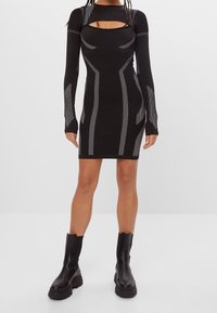 Musta bodycon-mekko pitkilla hihoilla, jossa on leikattu kaula-aukko ja hopeiset viivakoristeet. Yhdistetty mustien paksupohjaisten saappaiden kanssa.