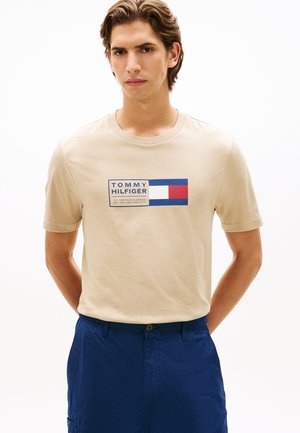 Mladi moški nosi bež majico Tommy Hilfiger z logotipom in modre hlače, stoji pred enobarvno belo ozadje.