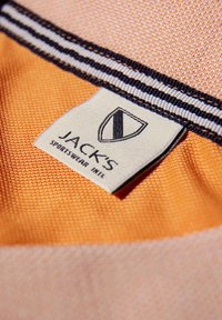 Pomarańczowy materiał z teksturowanym wykończeniem. Zawiera metkę z napisem "JACK'S" oraz logo w formie tarczy, uzupełnioną czarno-białym paskiem.