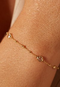 Gouden armband met gladde schakels, met twee ronde facettestenen die zijn ingebed in cirkelvormige zettingen langs de ketting. Textuur zichtbaar op de huid.
