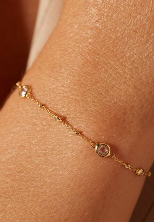 Gouden armband met gladde schakels, met twee ronde facettestenen die zijn ingebed in cirkelvormige zettingen langs de ketting. Textuur zichtbaar op de huid.
