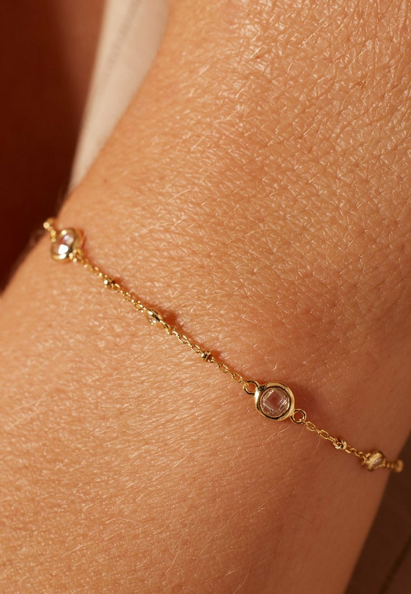 Gouden armband met gladde schakels, met twee ronde facettestenen die zijn ingebed in cirkelvormige zettingen langs de ketting. Textuur zichtbaar op de huid.